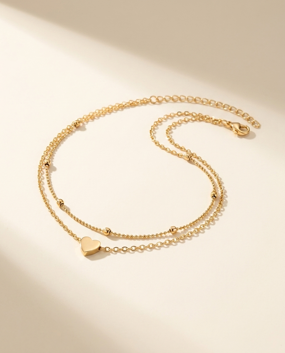 Gold double-chain heart anklet on a warm ivory studio background