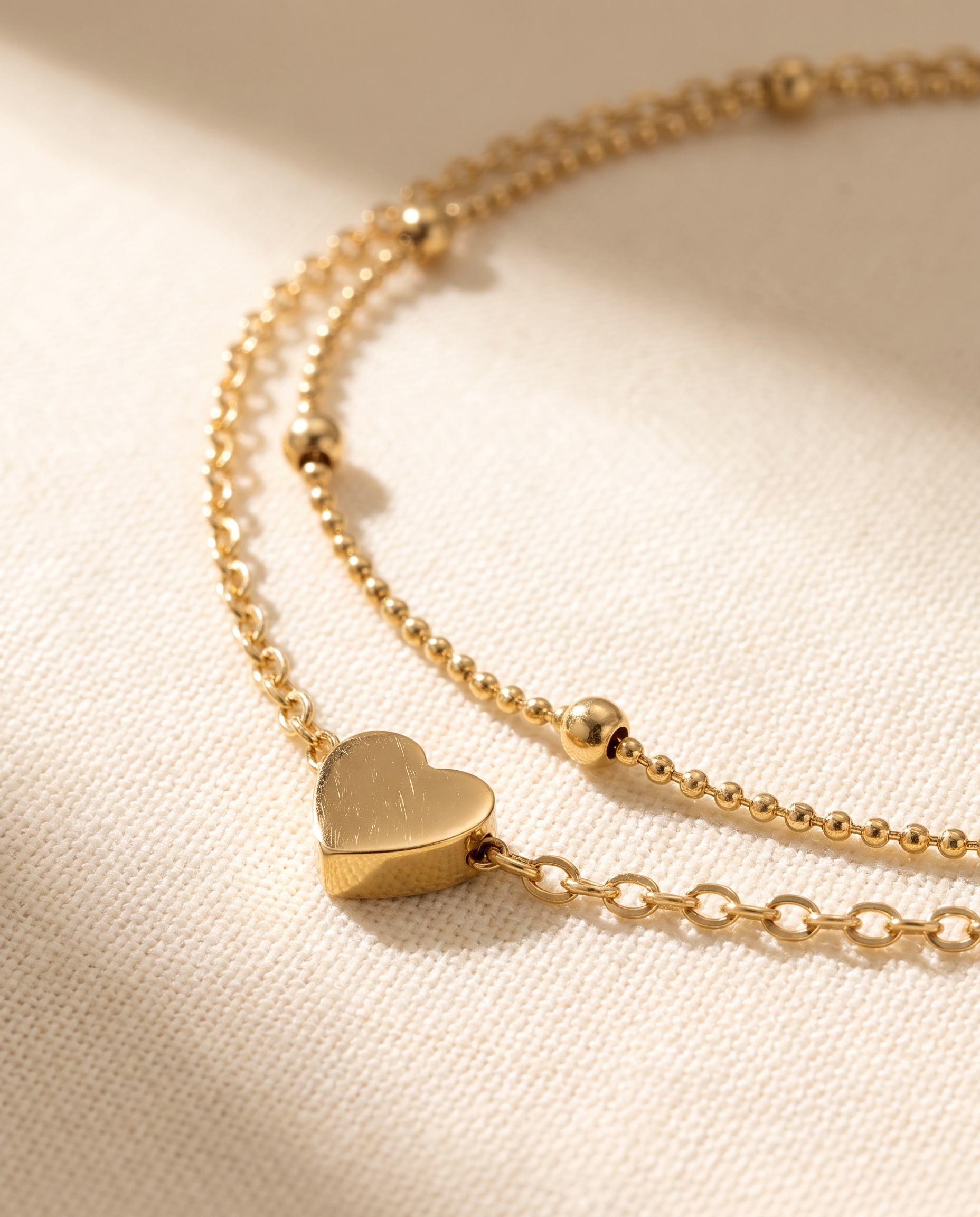 Close-up of the gold heart charm and double beaded chain of the anklet on warm ivory