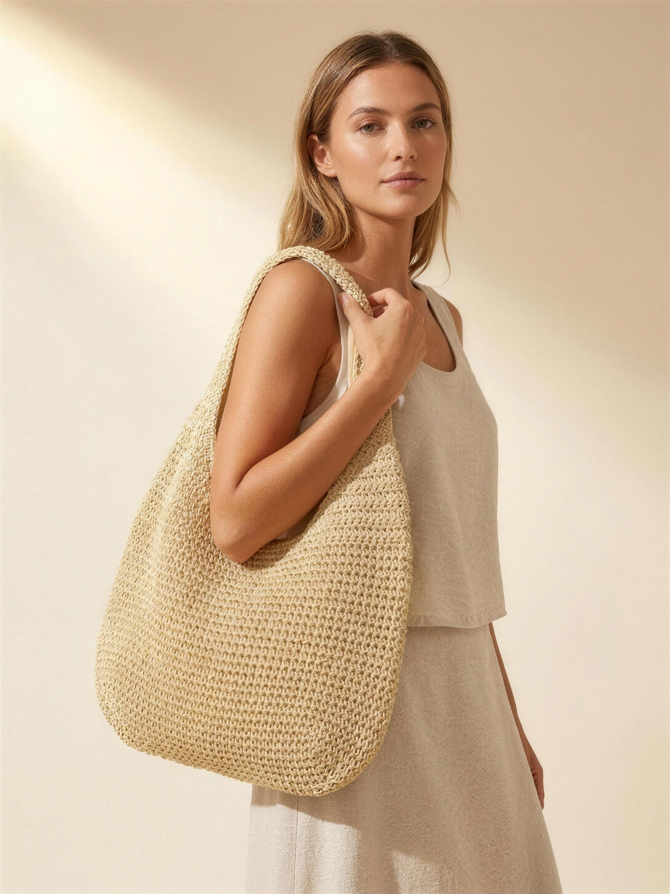 Model carrying the woven straw beach tote bag on her shoulder, warm ivory studio